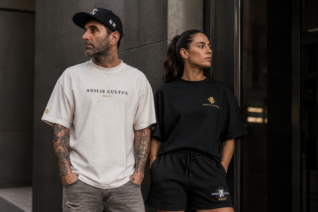 Man and woman wearing Agilis Cultus streetwear in black and bone with embroidered lion details against a modern urban wall.