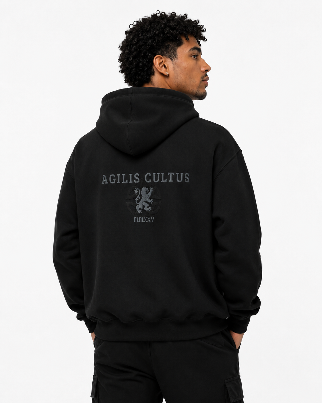 Male model wearing a black Agilis Cultus hoodie with subtle embroidered back logo and lion crest on a clean white studio background.