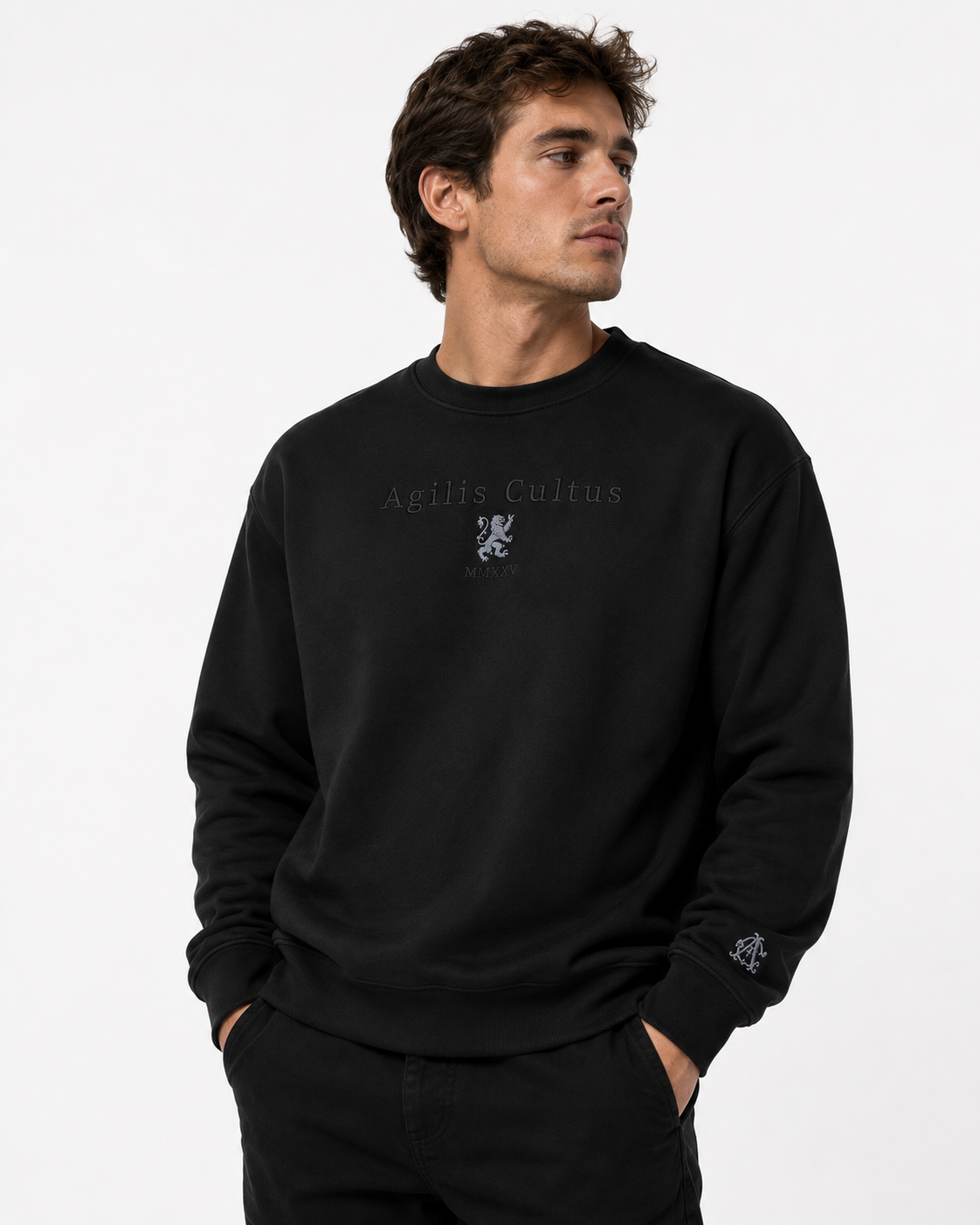 Male model wearing a black Agilis Cultus crewneck sweatshirt with subtle embroidered chest logo and sleeve monogram on a clean white studio background.