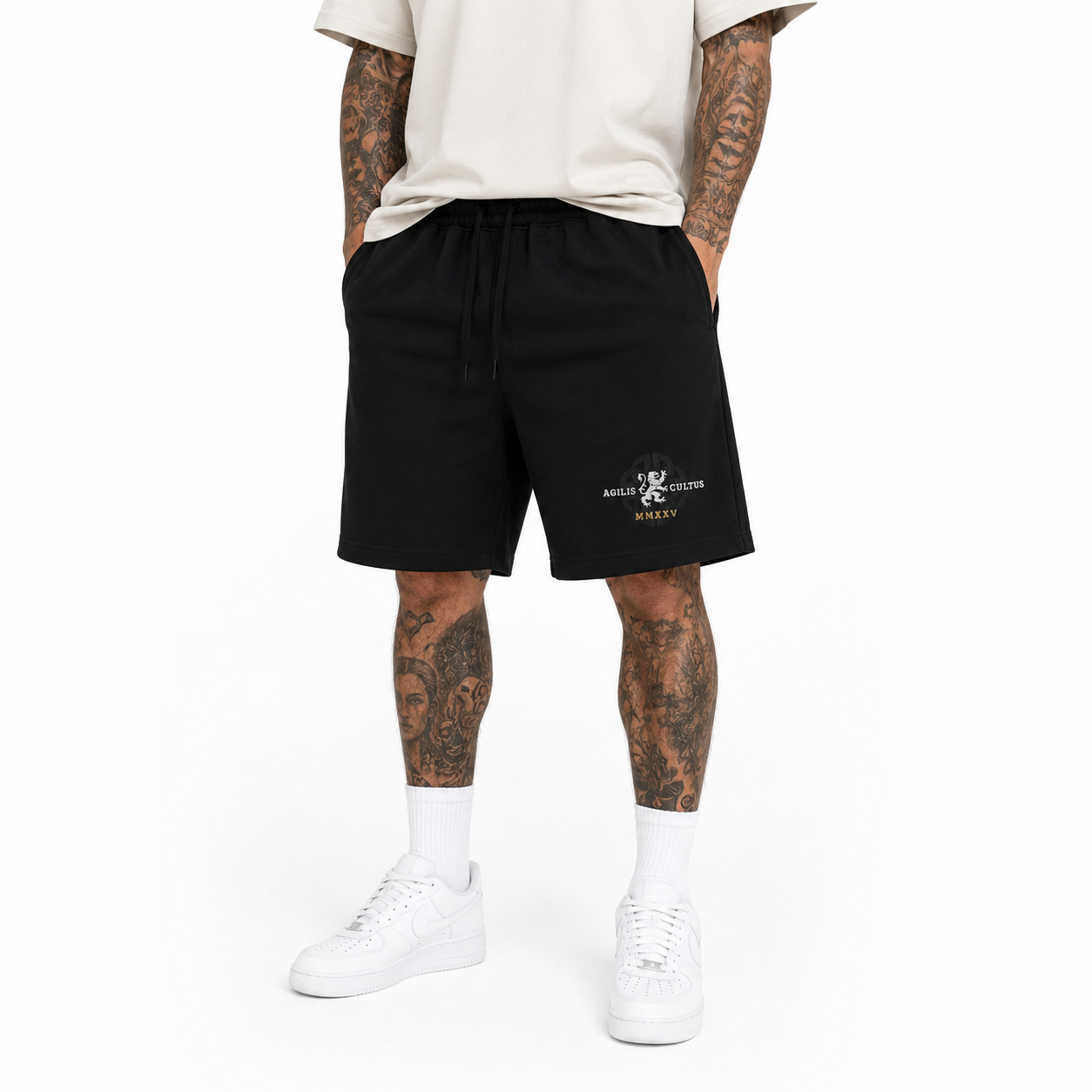 Male model wearing black Agilis Cultus fleece shorts with logo graphic, white t-shirt, white socks, and white sneakers on a clean studio background.