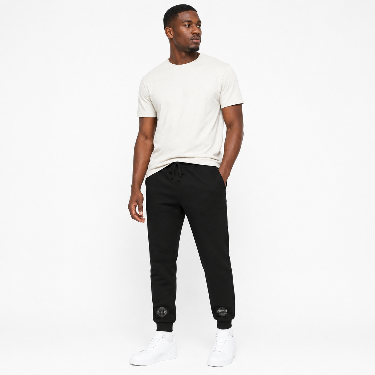 Male model wearing black Agilis Cultus joggers with ankle logo graphics, white t-shirt, and white sneakers on a clean white studio background.