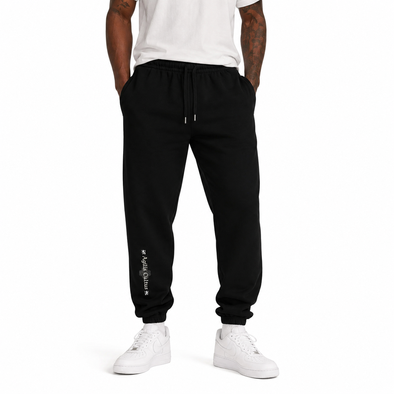 Male model wearing black Agilis Cultus sweatpants with vertical leg logo, white t-shirt, and white sneakers on a clean white studio background.