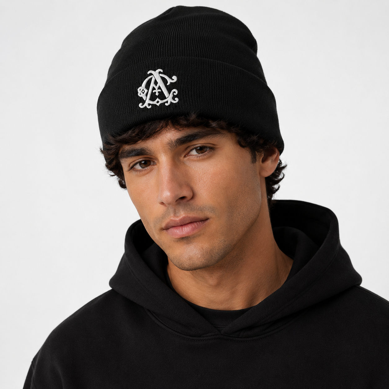 Male model wearing a black knit beanie with a white embroidered Agilis Cultus monogram on a clean white studio background.