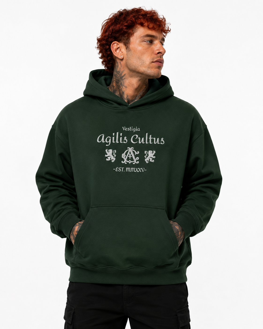 Red-haired male model wearing a dark green Agilis Cultus Vestigia hoodie with front lion and monogram artwork on a clean white studio background.