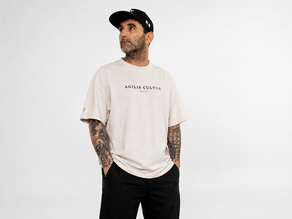 Tattooed male model wearing an oversized ivory Agilis Cultus t-shirt with black pants and a black cap on a clean white studio background.