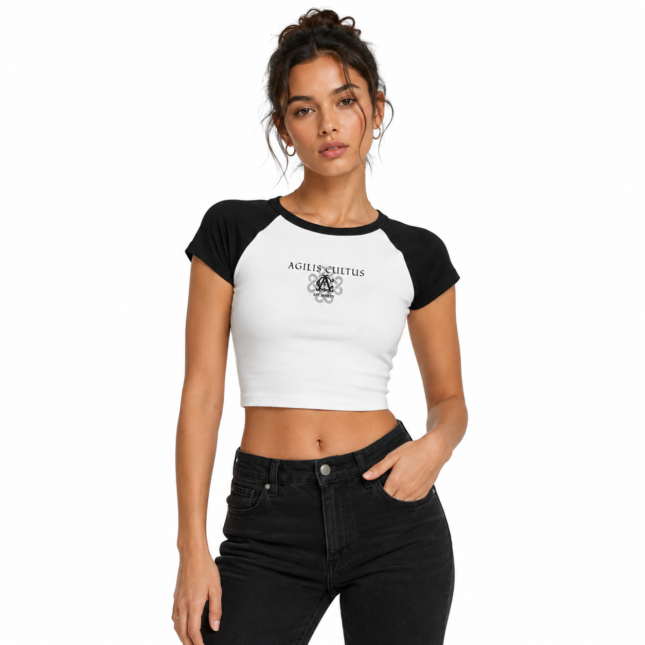 Female model wearing a fitted white and black Agilis Cultus micro baby raglan tee with front logo graphic and high-waisted black jeans on a clean white studio background.